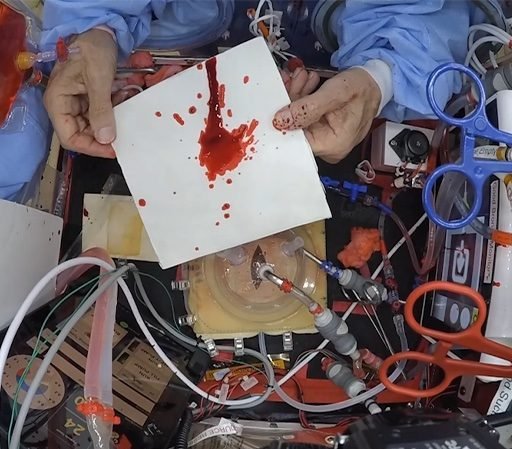 A cluttered workspace with electronic components and a hand holding a card splattered with red liquid.
