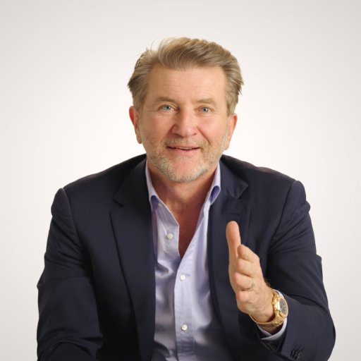A middle-aged man in a navy suit and light blue shirt gestures with his right hand while sitting against a plain light background.