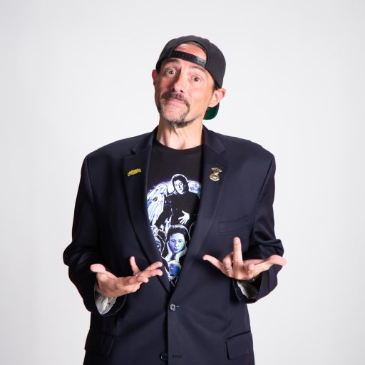 A man wearing a black blazer, graphic T-shirt, and backwards cap stands against a plain background with his hands raised in a questioning gesture.