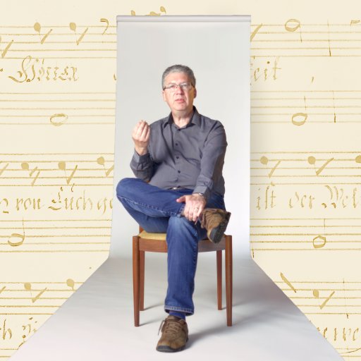 A man sits on a wooden chair with one leg crossed, gesturing with his hand, against a background of vintage sheet music.
