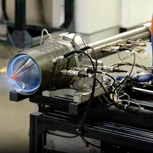 A close-up of a rocket engine testing setup, showing a small blue flame at the nozzle and multiple cables and pipes connected to the engine.
