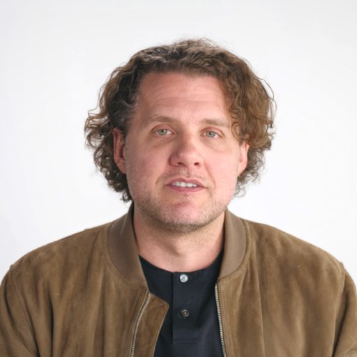 A man with wavy brown hair wearing a brown suede jacket over a black shirt sits in front of a plain white background.