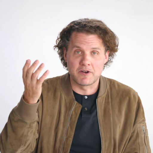 A man with curly hair wearing a brown suede jacket and black shirt gestures with his left hand while looking at the camera against a plain white background.