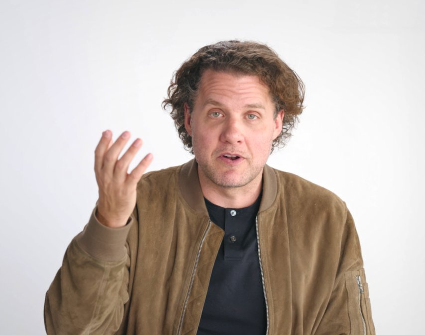A man with curly hair wearing a brown suede jacket and black shirt gestures with his left hand while looking at the camera against a plain white background.