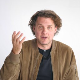A man with curly hair wearing a brown suede jacket and black shirt gestures with his left hand while looking at the camera against a plain white background.