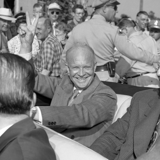 Two men smiling and riding in a convertible during a parade, surrounded by a crowd of onlookers.
