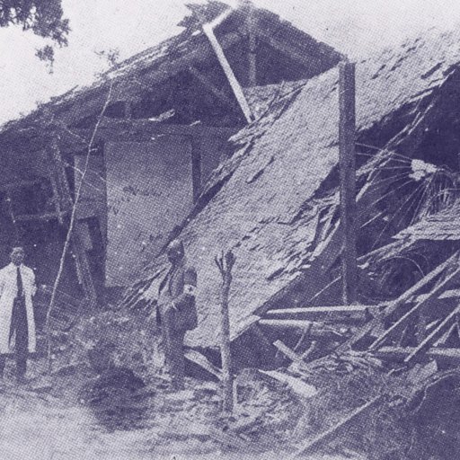 A man stands in front of a collapsed wooden building with debris scattered around in an outdoor setting.