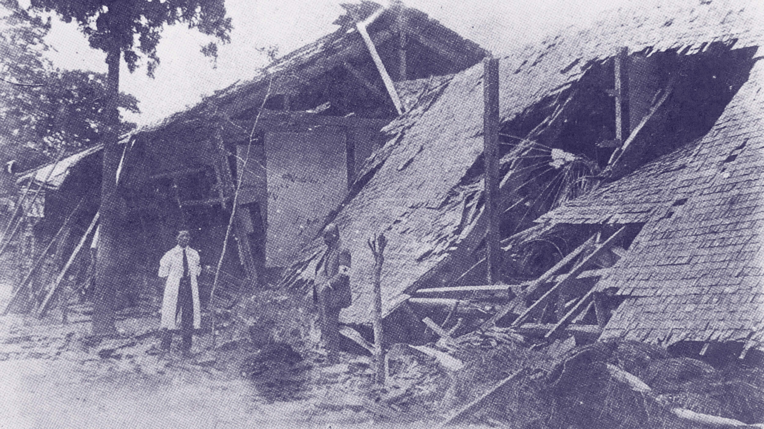 A man stands in front of a collapsed wooden building with debris scattered around in an outdoor setting.