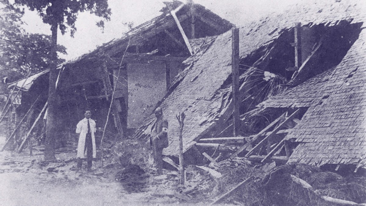 A man stands in front of a collapsed wooden building with debris scattered around in an outdoor setting.