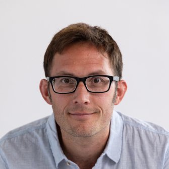 A man with short brown hair and glasses wearing a light blue button-up shirt looks directly at the camera against a plain white background.