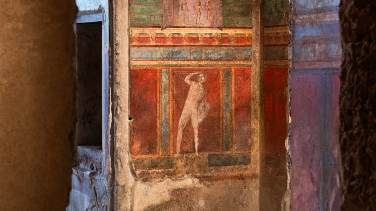 Ancient wall fresco depicting a standing human figure, surrounded by red, green, and brown decorative panels—an evocative remnant bearing the marks of history’s lost voices and the passage of time.