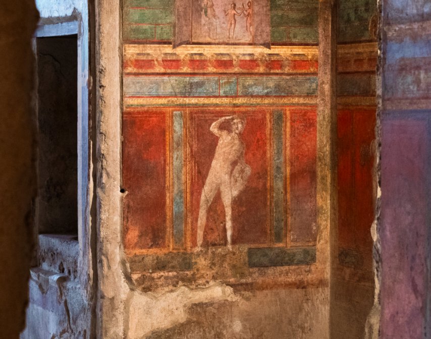 Ancient wall fresco depicting a standing human figure, surrounded by red, green, and brown decorative panels—an evocative remnant bearing the marks of history’s lost voices and the passage of time.