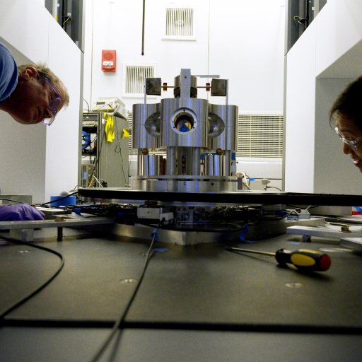 Two people examine scientific equipment in a laboratory, working on a large metallic device surrounded by cables and tools, as they investigate the physics mystery G—the elusive mystery gravitational constant.