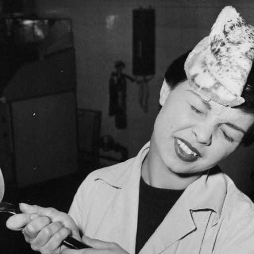 A woman displays stoicism as she grimaces, unfazed by a pancake landing on her head while holding a frying pan in a kitchen setting.