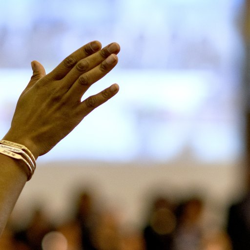 A raised hand with bracelets is shown in focus against a blurred background of people indoors.