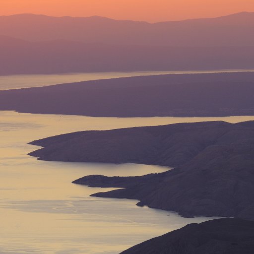 A coastal landscape with rugged cliffs shaped by seaflooding and calm water at sunset, with mountains in the background and soft orange and purple hues in the sky.
