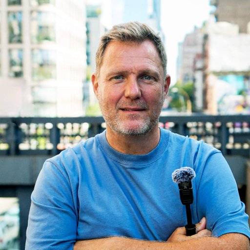 Simon Squibb, a middle-aged man in a blue shirt, stands outdoors in an urban setting, holding a small microphone with his arms crossed.