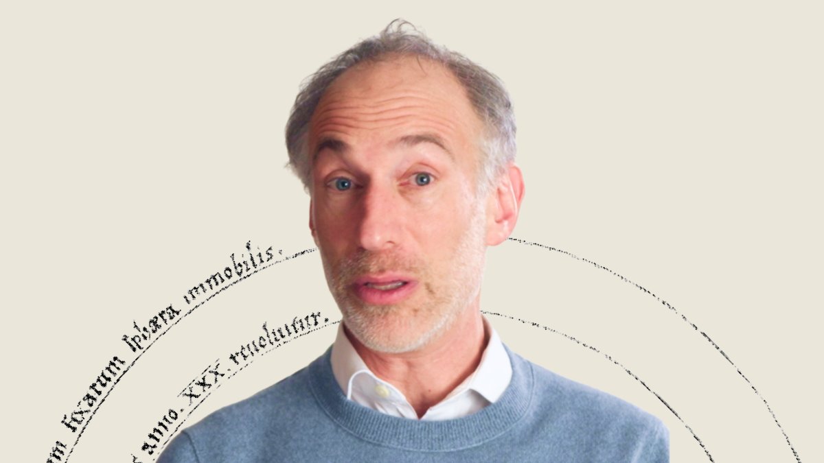 A middle-aged man with light hair and a beard, wearing a blue sweater over a white shirt, stands in front of a neutral background with faint, curved text elements.