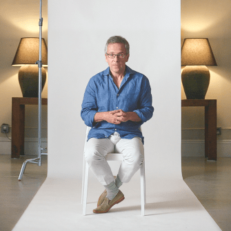 A man in a blue shirt and white pants sits on a white chair in a studio, with lamps and tables in the background and a white backdrop behind him.