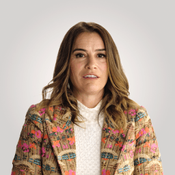 A woman with long brown hair wearing a patterned jacket and white textured top, facing forward against a plain light gray background.