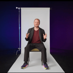 A person sits on a chair in front of a white backdrop, gesturing with both hands. The background fades from dark blue to purple. The BT logo appears in the top right corner.